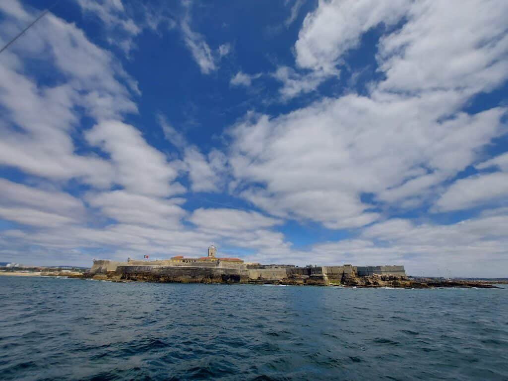 Vista desde o mar sobre o Forte de S. Julião da Barra, com um céu azul e nuvens brancas