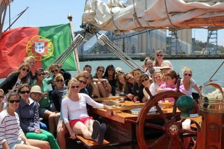 A bandeira portuguesa está visível, pendurada na parte traseira do barco, enquanto os convidados aproveitam o ambiente descontraído e a vista do rio.