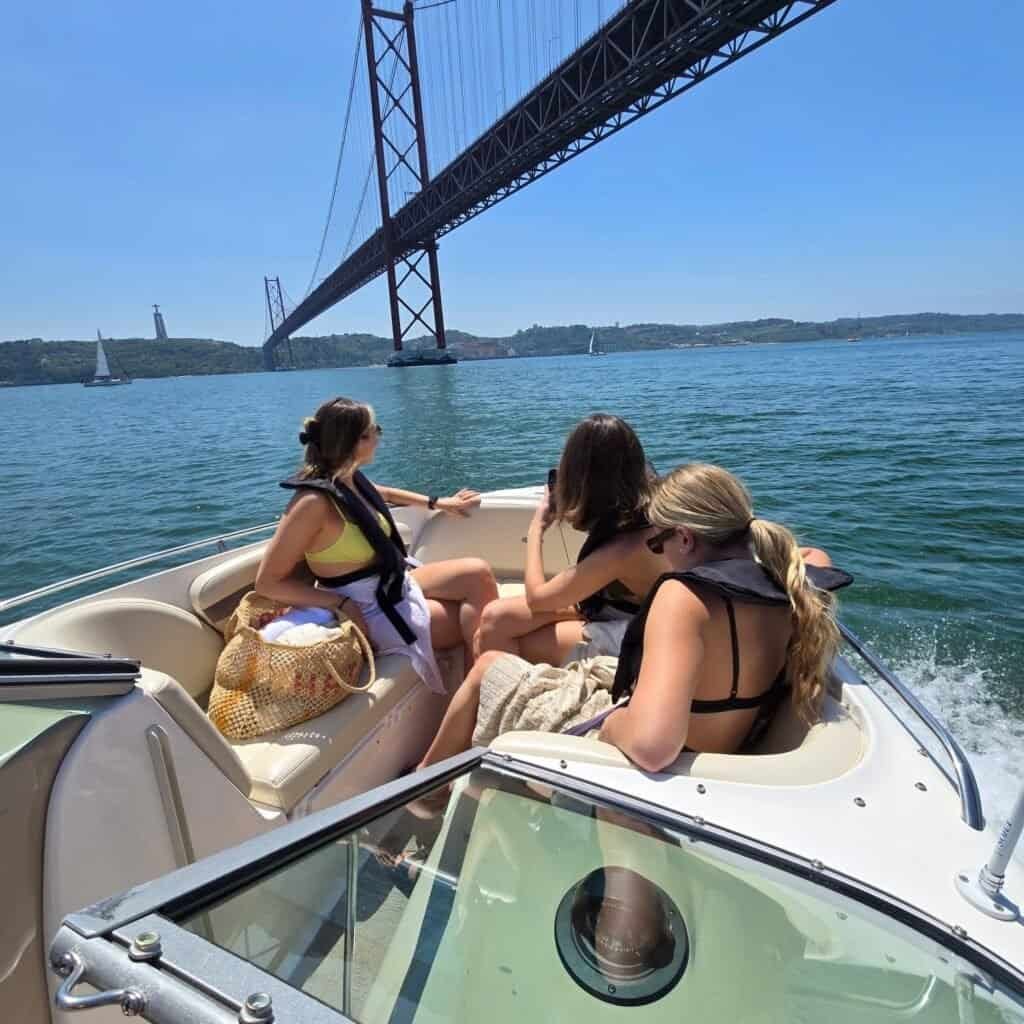 Group of friends on board the yacht, sailing under the 25 de Abril Bridge with a view of Cristo Rei.