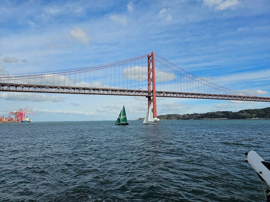 A foto capta a ponte com a sua característica estrutura vermelha contra um céu azul com nuvens. Em primeiro plano, o rio está agitado e há dois veleiros navegando sob a ponte, sugerindo o seu uso contemporâneo e turístico, além de ser um monumento reconhecido de Portugal.