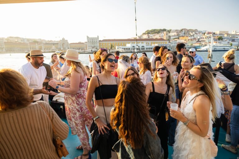 Shows a group of people socializing under the sunset light, with the Monument to the Discoveries or the 25th of April Bridge in the background.