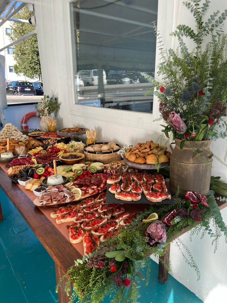 A colorful and bountiful buffet table, decorated with rustic floral arrangements, featuring a variety of cheeses, sausages, fruits, and canapés