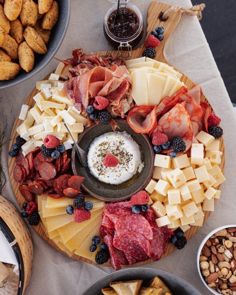 Apresenta uma seleção de queijos curados em cubos, presunto fatiado, salame e frutos vermelhos. No centro, uma taça com queijo fresco decorado com ervas, tudo servido sobre uma mesa de madeira rústica, ideal para o início de um evento.