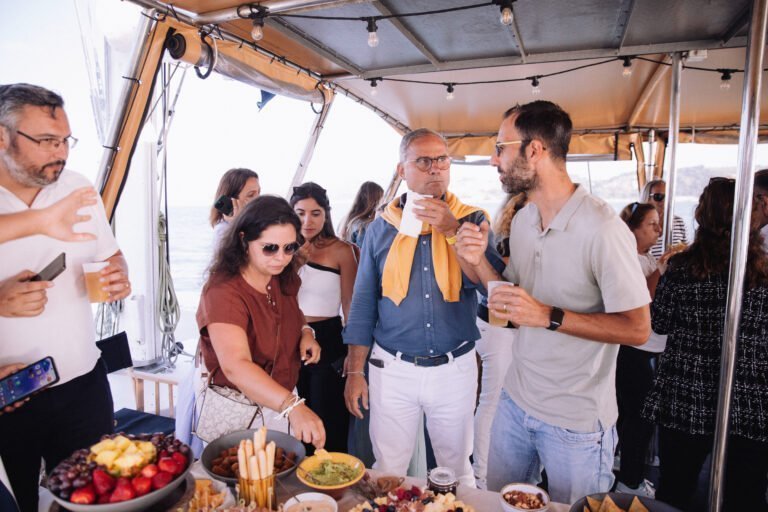 Convidados em traje casual-chique conversam enquanto seguram bebidas, aproveitando o ambiente social. Ao fundo, vê-se a estrutura moderna do barco com proteções laterais transparentes que permitem ver o rio.