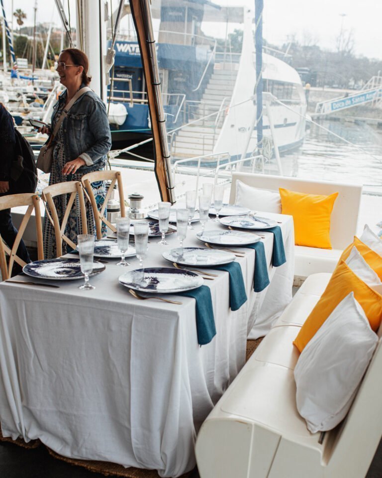 A configuração de jantar formal no convés. Uma mesa comprida vestida com uma toalha branca impecável, preparada com pratos decorativos em tons de azul e branco, talheres e copos de vinho.