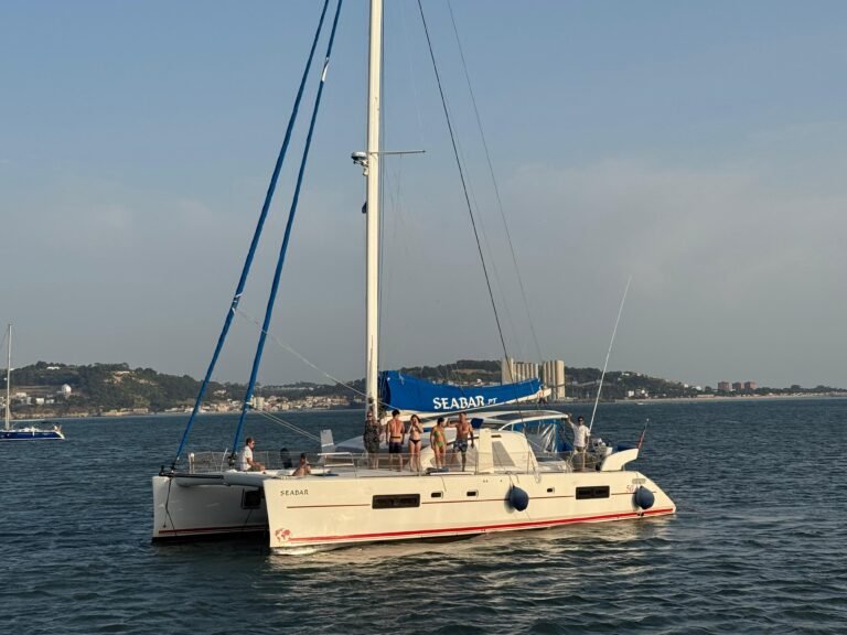 Photograph of a modern white catamaran sailing on the Tagus River, in Lisbon. The boat has 'SEABAR' written on the folded sail and on the hull. Several people are on the deck enjoying the tour. In the background, you can see the Lisbon hillside with vegetation and buildings under a clear late-afternoon sky