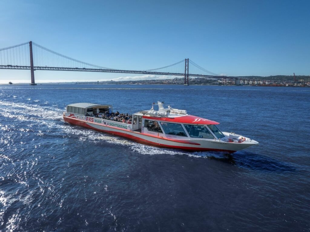 An exterior view of the motor vessel Hanseblick sailing on the River Tagus, with the 25 de Abril Bridge in the background. A 122-foot vessel ideal for large events in Lisbon.