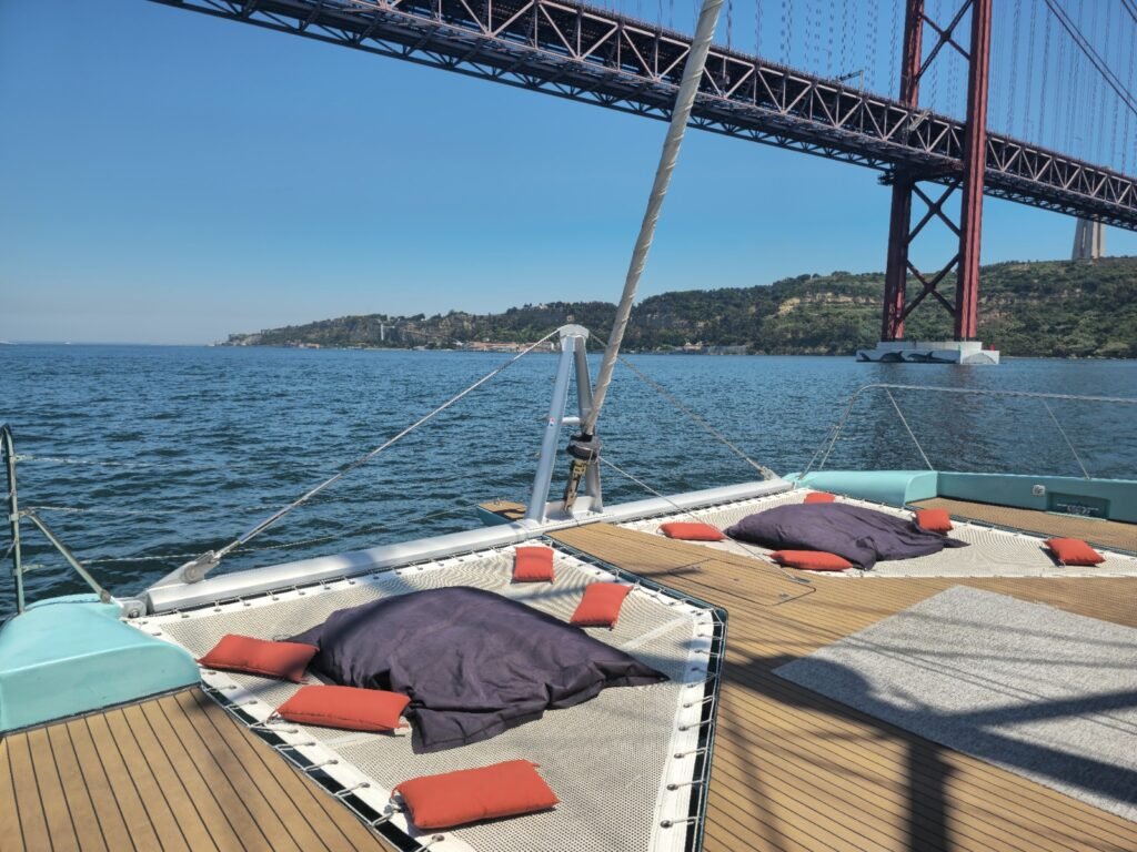 La proa de un catamarán con cojines de colores y hamacas, con el puente 25 de Abril al fondo.
