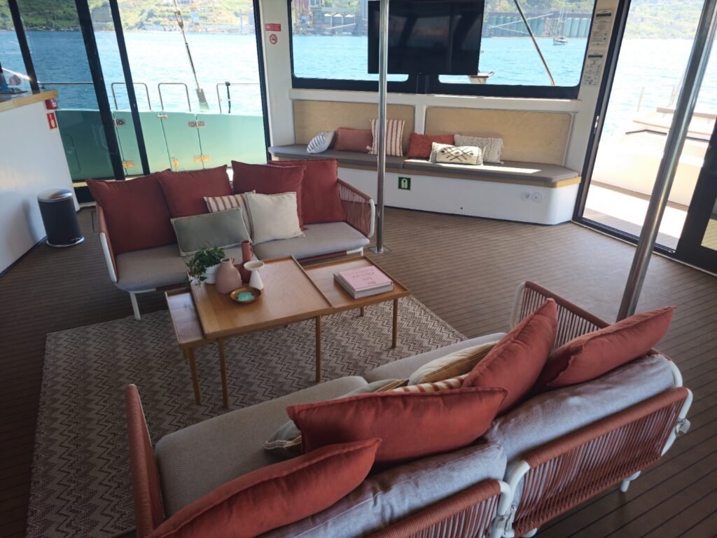 Salón interior de un catamarán con sofás grises y cojines rojos, grandes ventanas panorámicas y un televisor.