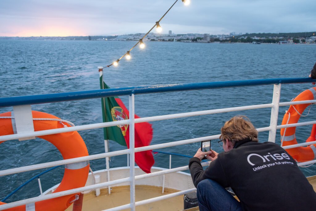 Um dos colaboradores da empresa, com o casaco oficial da ORISE, fotografa a bandeira portuguesa à popa do barco durante o entardecer no Tejo.