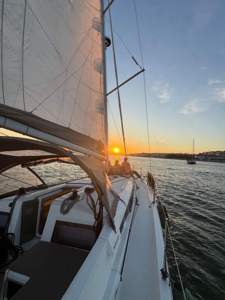Pôr do sol visto do convés do veleiro Maharani, com o sol a tocar a água e reflexos alaranjados. Passeio privado ao pôr do sol em Lisboa.