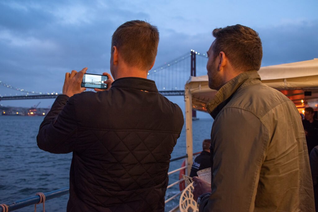 Invitados de ORISE capturan fotografías del Puente 25 de Abril desde la cubierta superior del barco Évora, deslumbrados por la belleza de Lisboa vista desde el río.