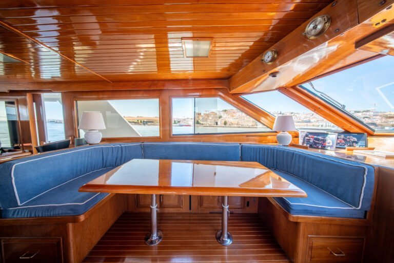 Coin salon avec un canapé en U recouvert de tissu bleu, une table en bois et une vue panoramique à travers le pare-brise du bateau.