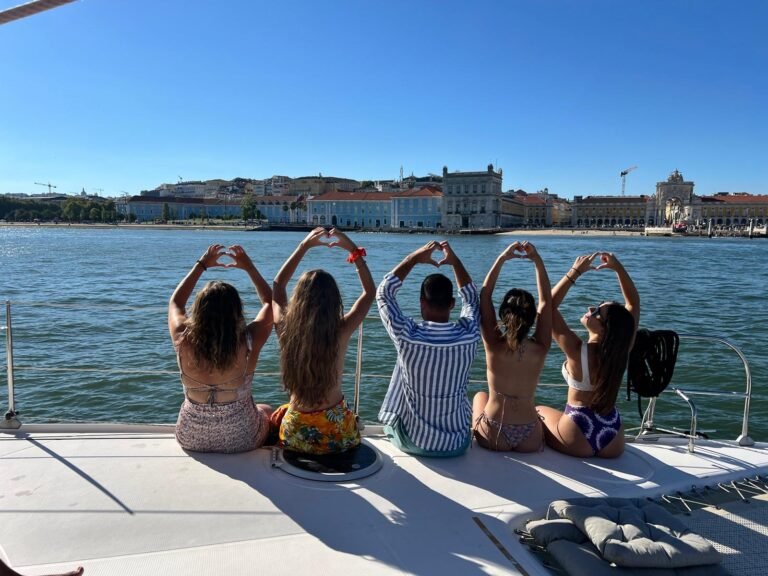 Grupo de amigos a bordo do catamarã Corto Maltese III, na proa do barco, com o rio Tejo e a zona ribeirinha de Lisboa em pano de fundo. Passeio de catamarã ideal para grupos, despedidas de solteira ou celebrações em Lisboa.