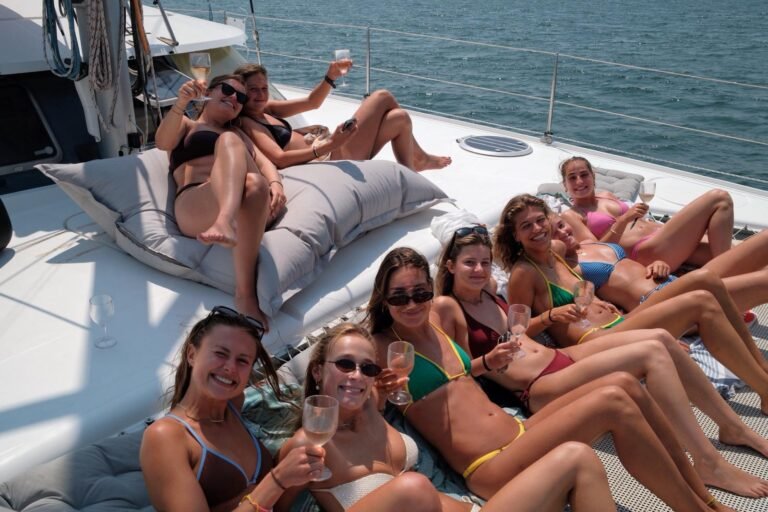 Grupo de amigas em ambiente de festa a bordo do catamarã Corto Maltese III, sentadas na proa do barco a brindar com bebidas, rodeadas pelo rio Tejo. Passeio de catamarã para despedidas de solteira, aniversários e festas privadas em Lisboa, com espaço espaçoso para grupos até 10 pessoas.
