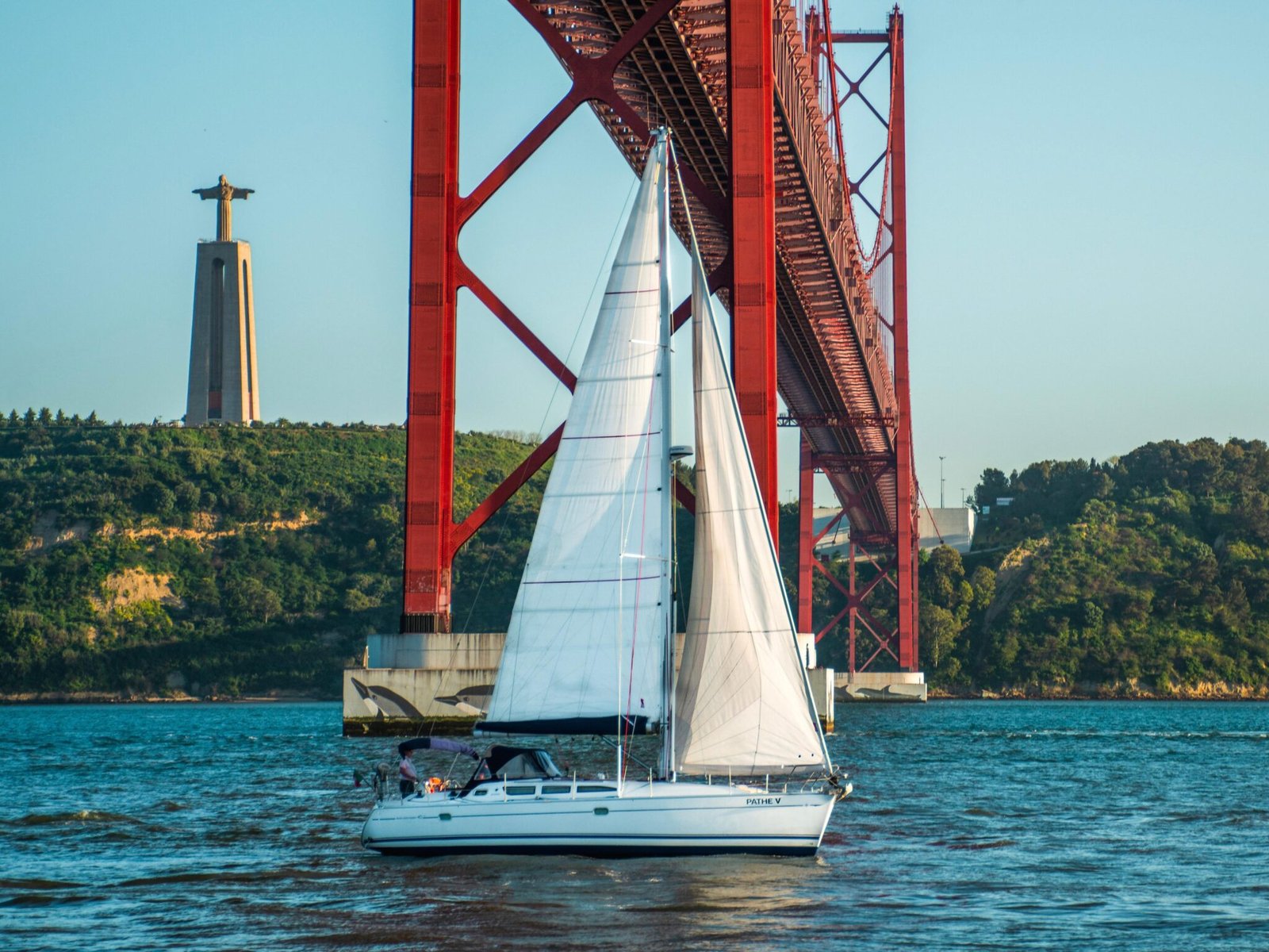 Veleiro a navegar no Rio Tejo junto à Ponte 25 de Abril, com o Cristo Rei ao fundo, em Lisboa